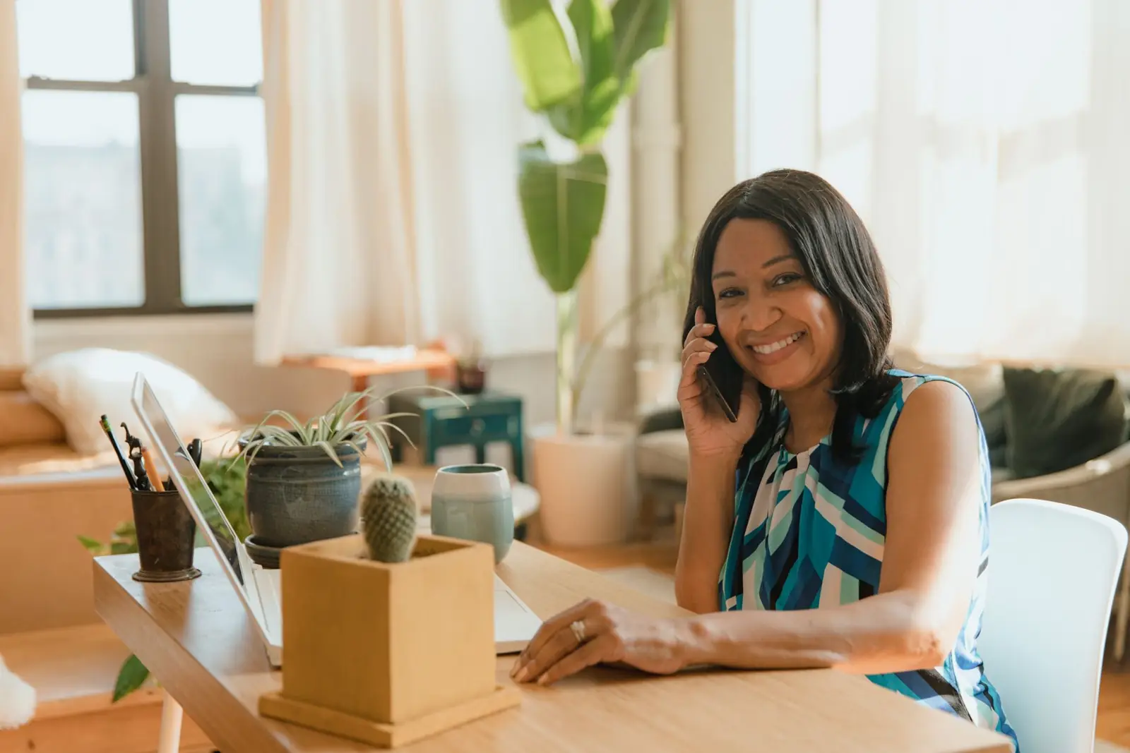 Smiling woman in home office talking on phone with laptop and plants on desk.